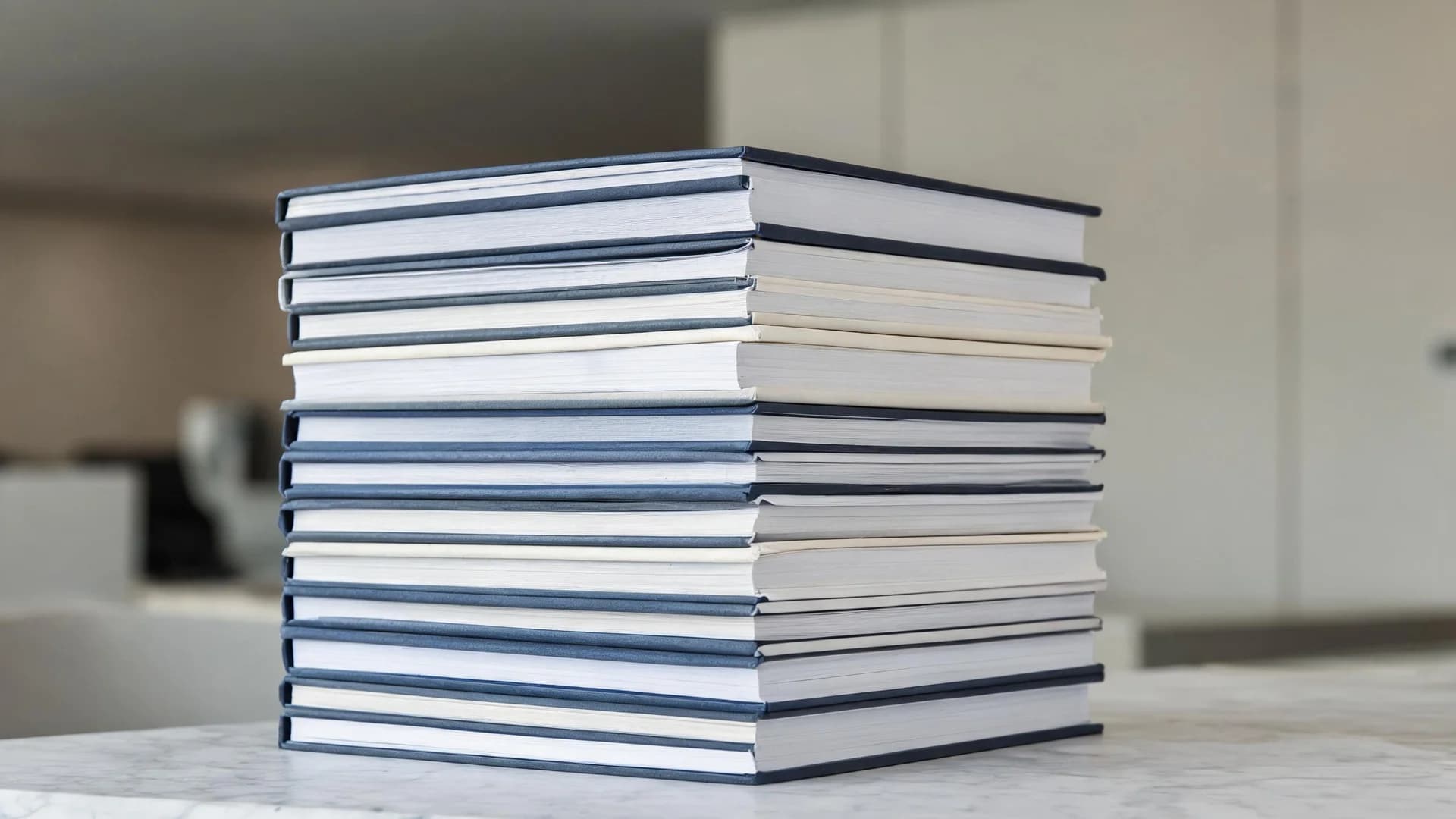A tall stack of thick bound corporate reports with plain unmarked covers standing on a pale marble surface, photographed from a low angle in soft daylight.