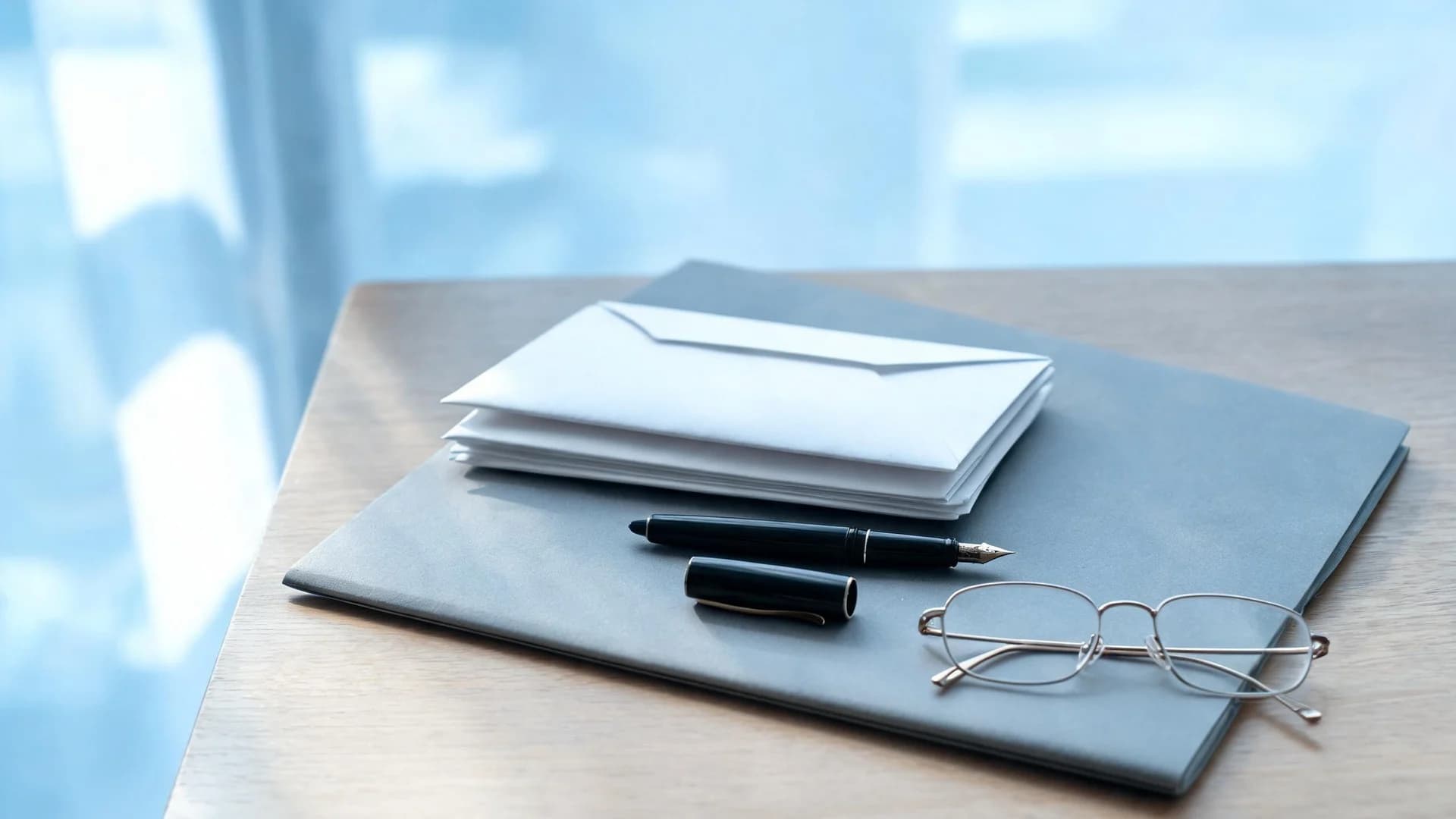 A stack of plain sealed envelopes, an uncapped fountain pen and reading glasses on a closed grey document file by a window