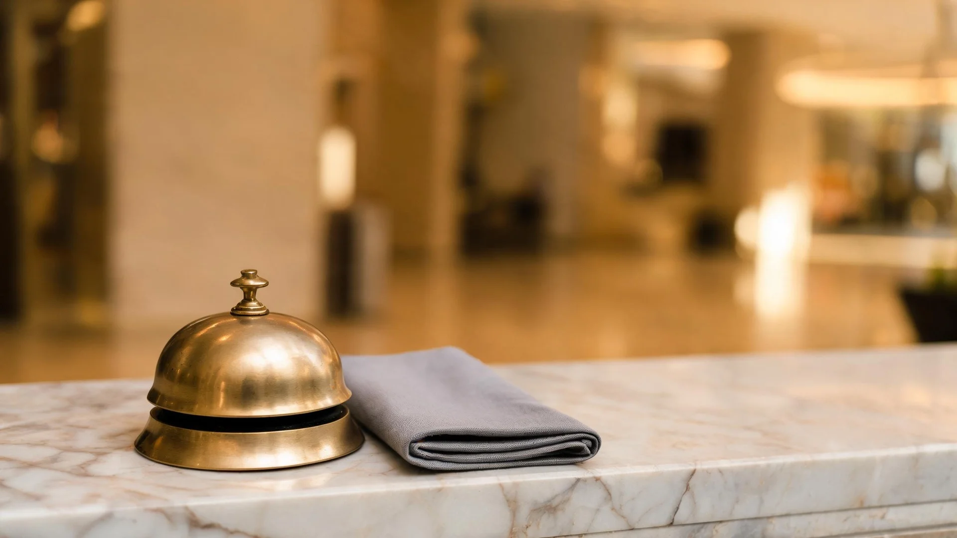 A polished brass counter bell standing on a pale marble ledge beside a folded grey cloth, with a quiet building interior softly blurred behind it in warm afternoon light.