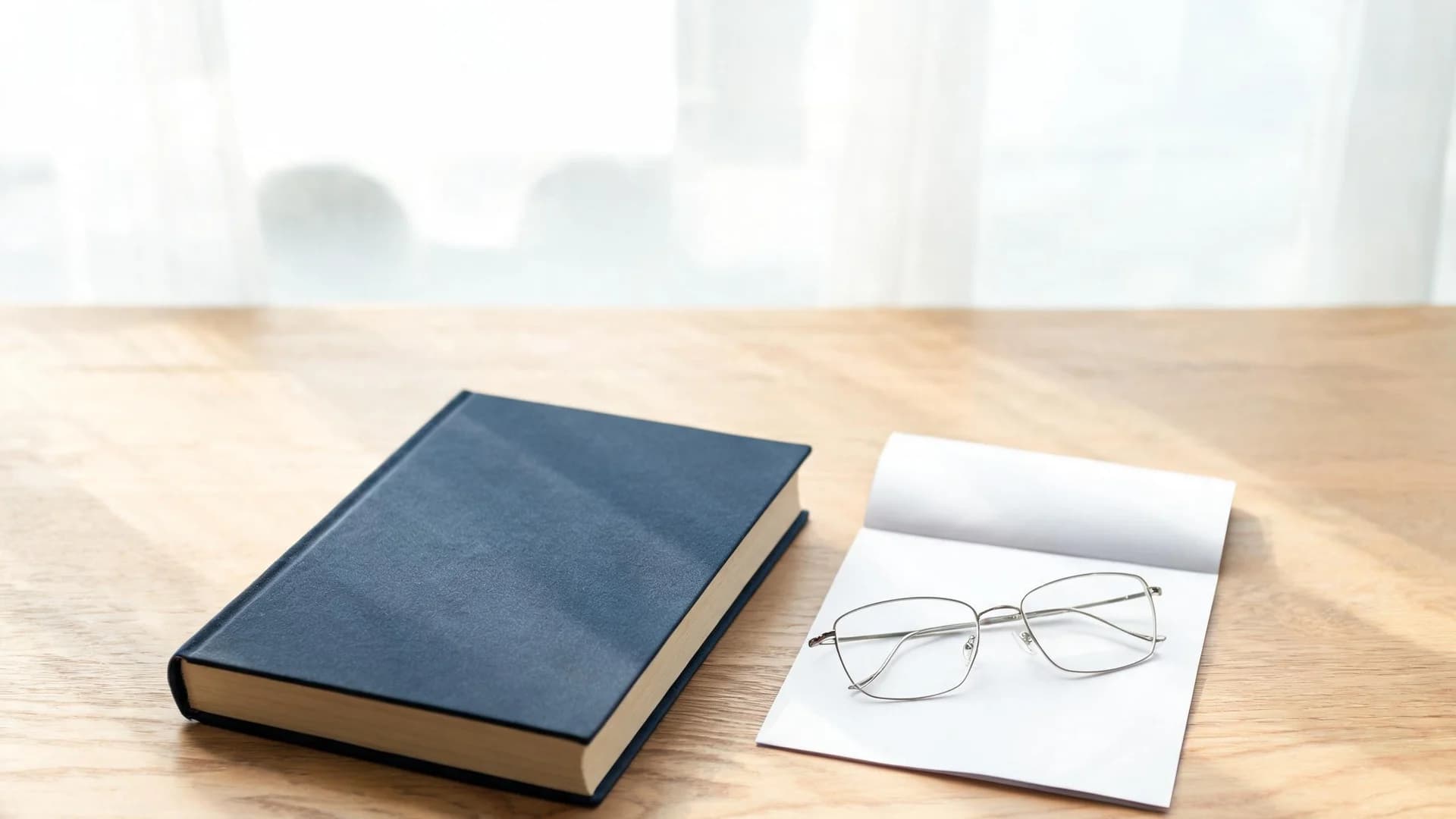 A thick navy hardbound book beside a single folded sheet of blank paper with reading glasses resting on it, on a light oak desk