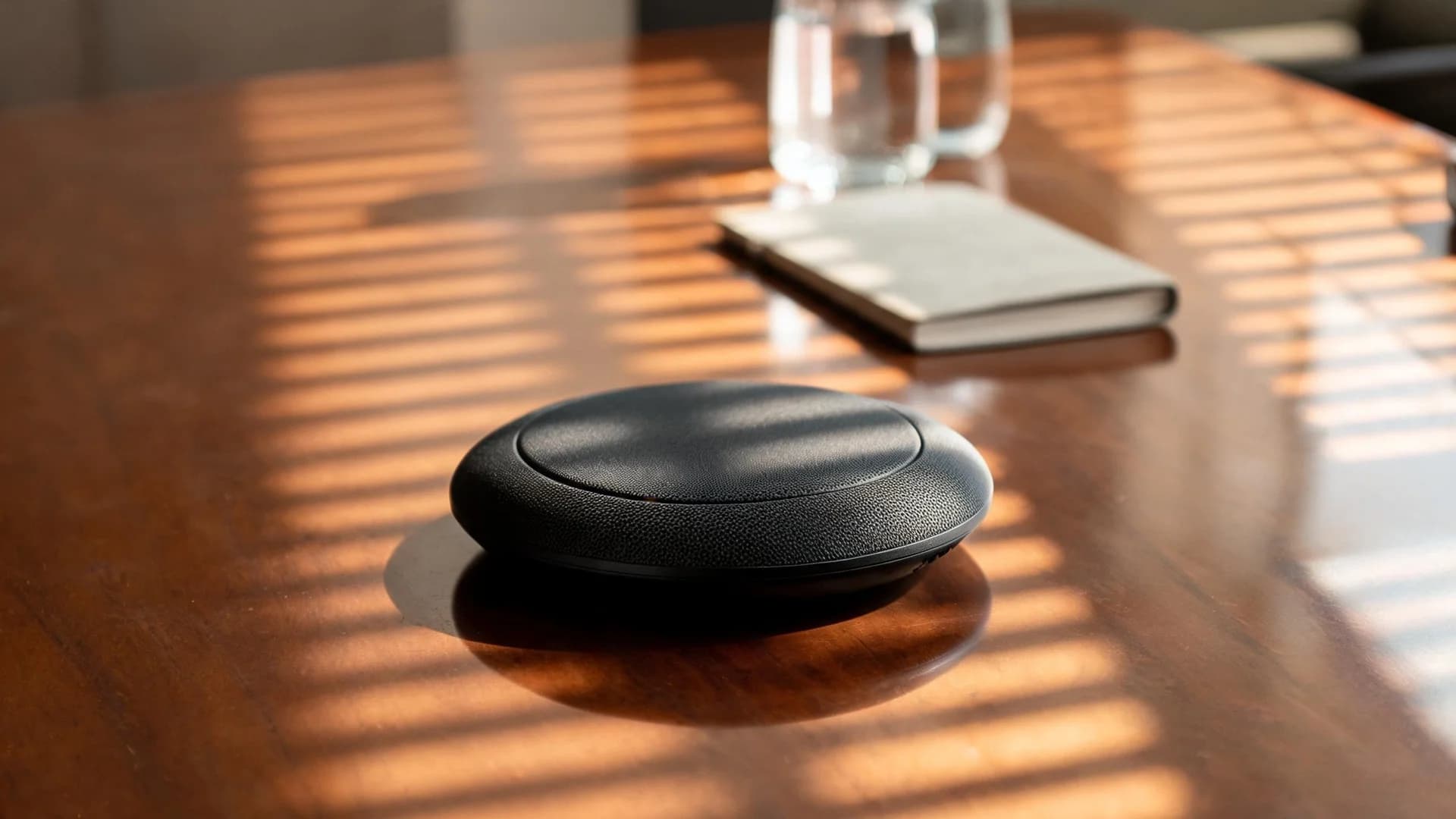 A black circular conference speakerphone on a polished boardroom table beside a closed bound booklet, with afternoon light falling through window blinds.