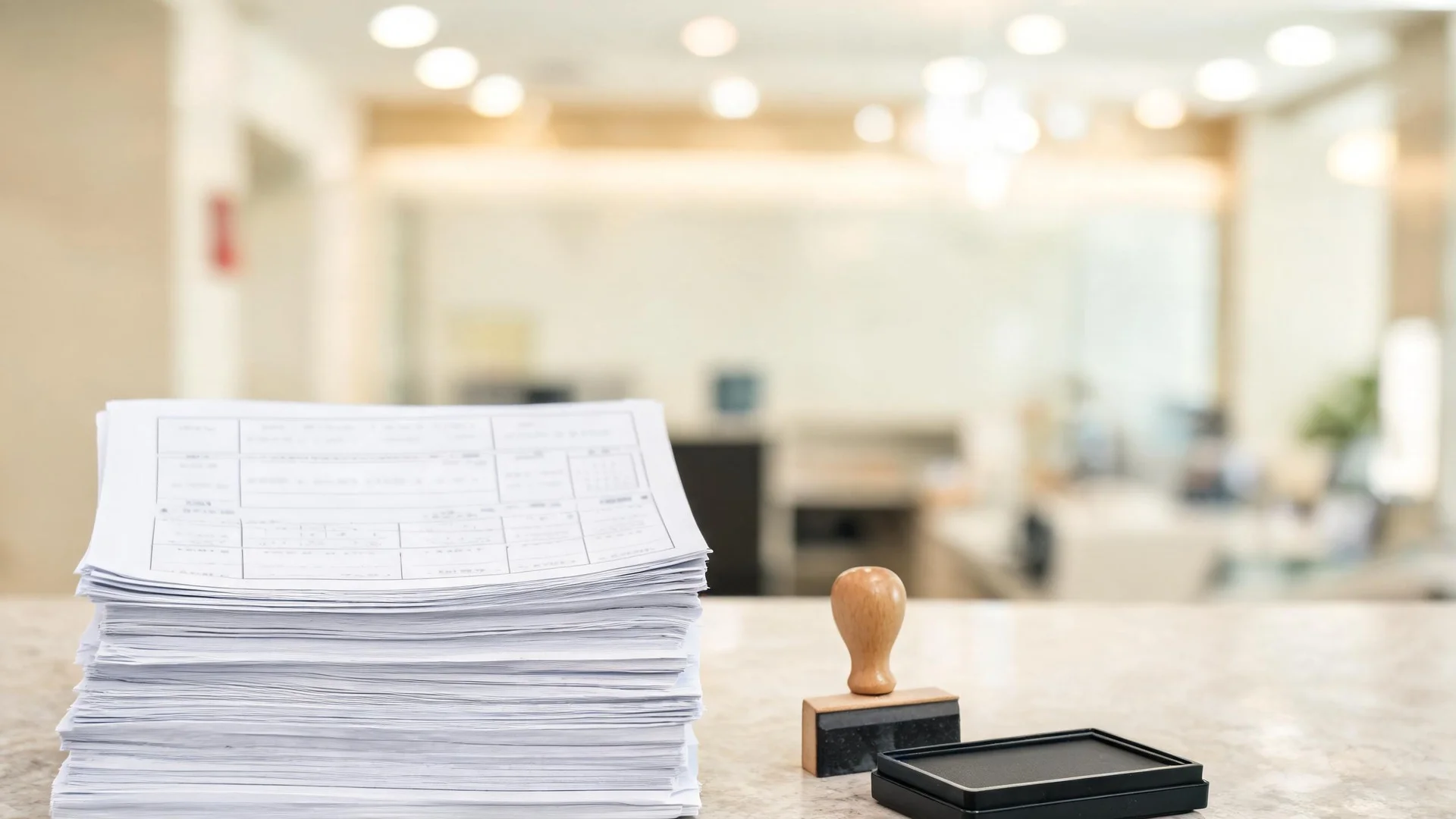 A tall stack of printed application forms on a pale stone counter with a wooden-handled rubber stamp and a flat ink pad beside them, in a softly blurred bright hall.
