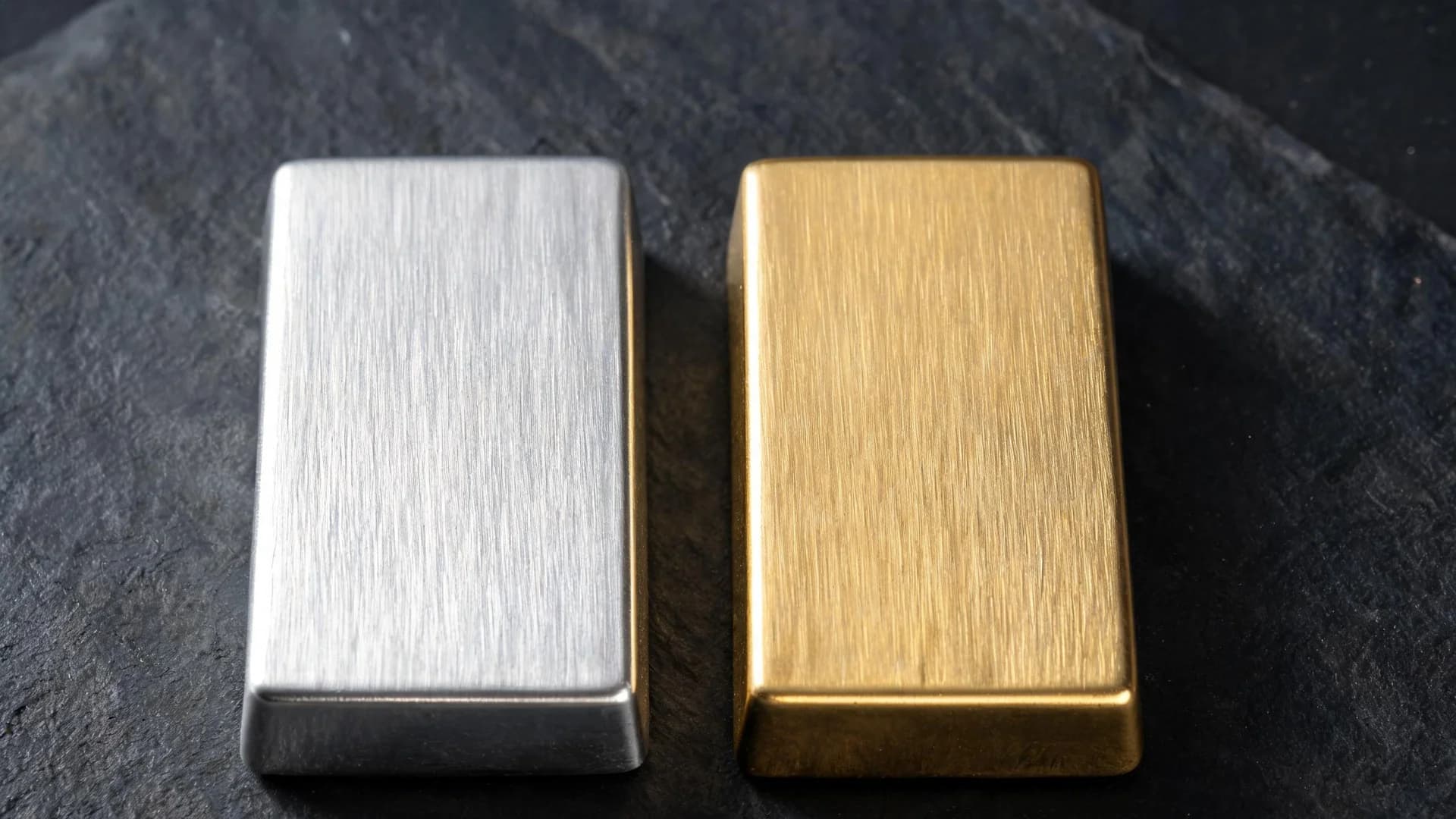 A plain silver bar and a plain gold bar of identical size and shape lying side by side on dark slate.